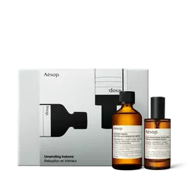 Zwei bernsteinfarbene Aesop-Flaschen mit schwarzen Verschlussen stehen vor einer silberfarbenen Schachtel, die mit schwarzen Flaschenillustrationen verziert ist.