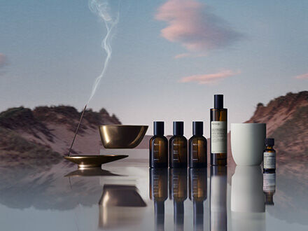 Eine Kollektion von Aesop-Duftprodukten fur den Hause, darunter bernsteinfarbene Glasspruhflaschen, ein Olbrenner und Weihrauch, die auf einer reflektierenden Oberflache vor einer ruhigen Wustenlandschaft bei Einbruch der Dammerung angeordnet sind.