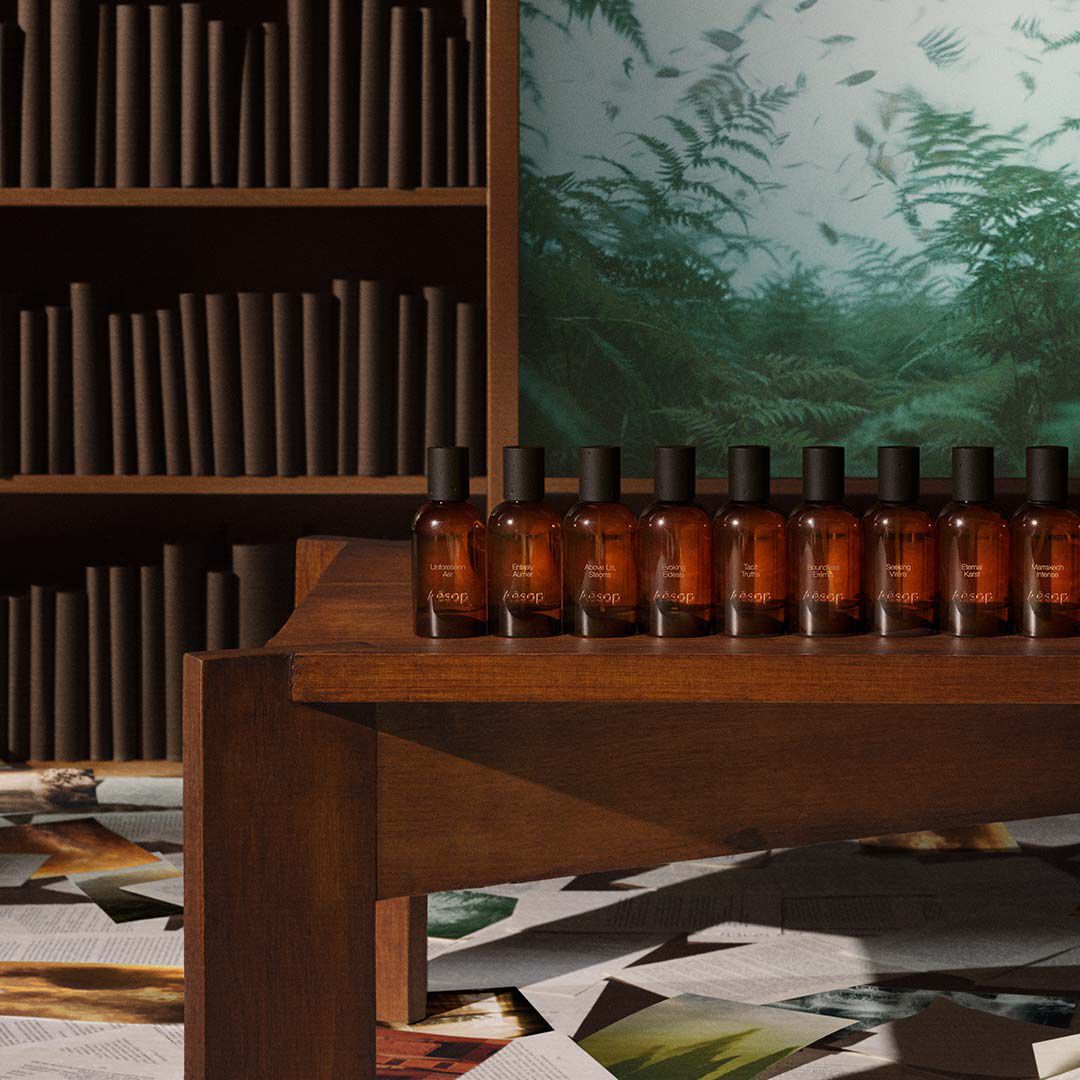 Eine Reihe von Aesop-Parfumflaschchen aus bernsteinfarbenem Glas auf einem Holzregal vor einer Bibliothek und einem grunen Kunstwerk.