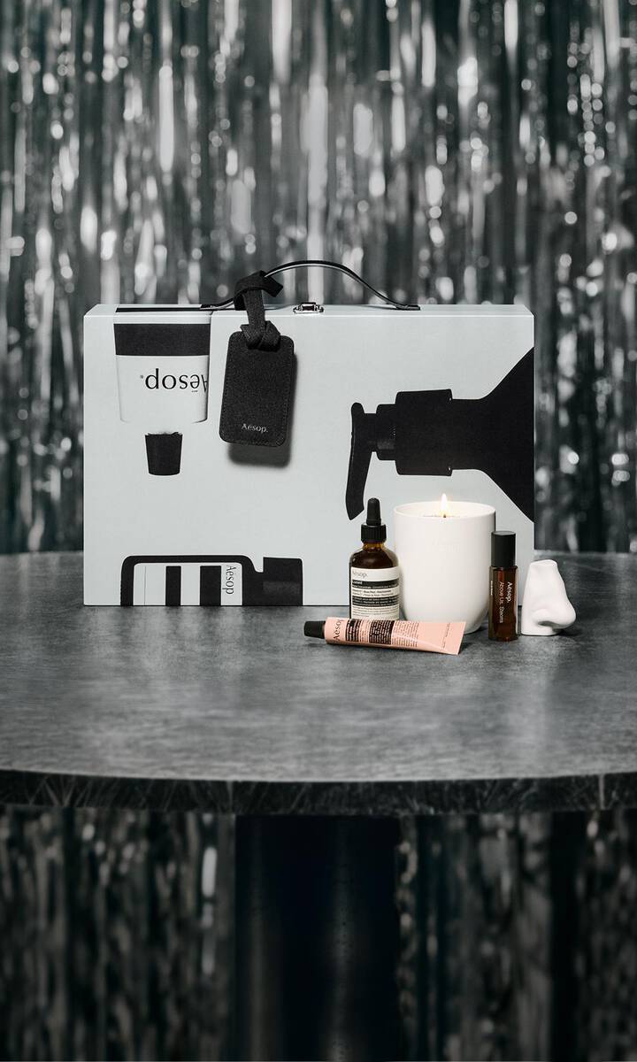 Aesop-Geschenkset und Hautpflegeprodukte auf einem Tisch vor festlich beleuchtetem Hintergrund.