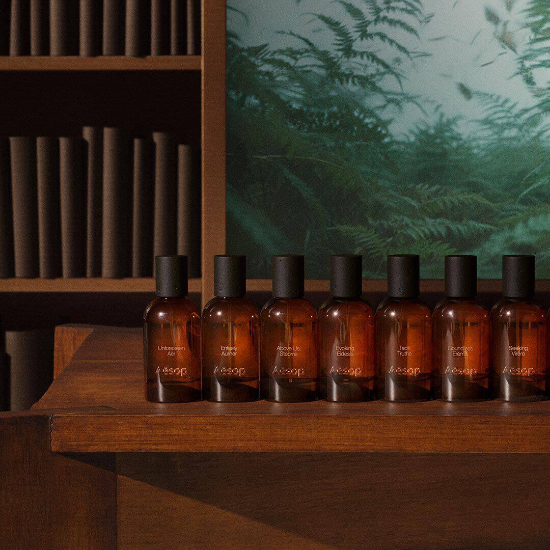 Reihe bernsteinfarbener Aesop-Duftflakons auf einem Holztisch neben Regalen und dem Bild eines grunen Farns.