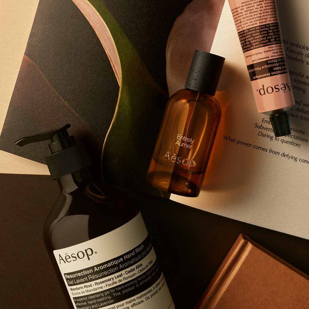 Aesop Entirely Aurner Flakon mit Pumpspender und pfirsichfarbenem Schlauch, arrangiert auf Papieren und geschwungenen dunklen Formen.