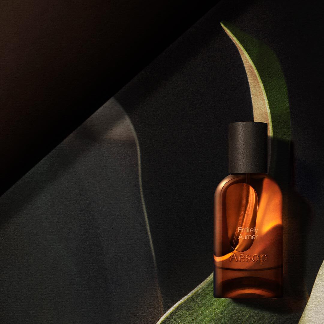 Einzelne bernsteinfarbene Aesop-Flasche, in warmes Licht getaucht, gerahmt von geschwungenen, blattartigen Formen in Dunkelgrun, Creme und Schwarz.