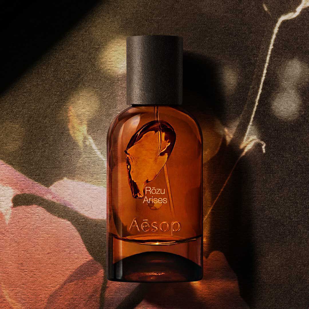 Bernsteinfarbener Aesop Rozu Arises Duftflakon auf einem dunklen floralen Bild mit warmem Licht und Schatten.