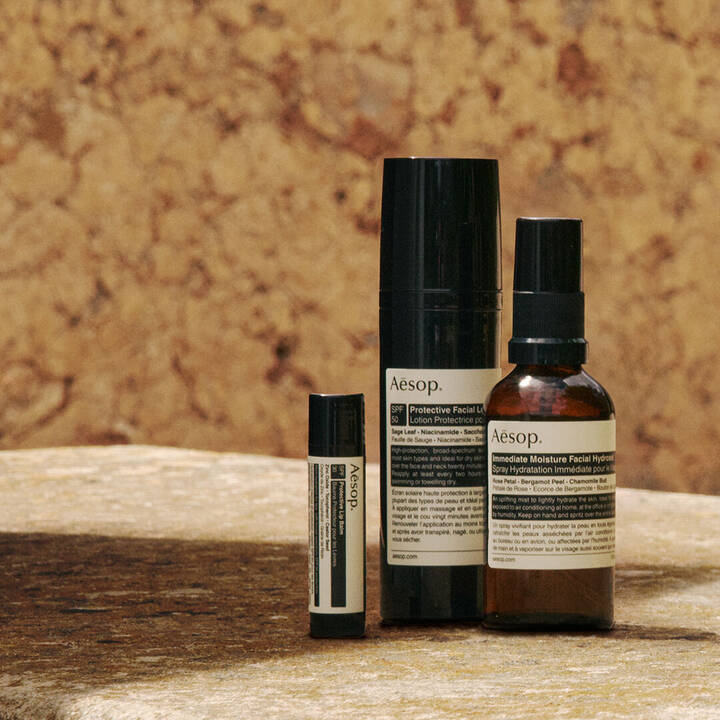 Eine sorgfaltige Auswahl an Aesop Sonnenschutz-Formulierungen, arrangiert auf einer sonnenbeschienenen Steinoberflache vor einer Wand in warmen Erdtonen.
