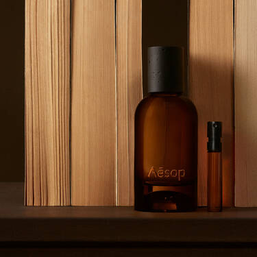 Bernsteinfarbene Aesop-Flasche und schmales Flaschchen zwischen vertikalen, hellen Holzbrettern, mit tiefbraunen Schatten und einer klaren, linearen Komposition.