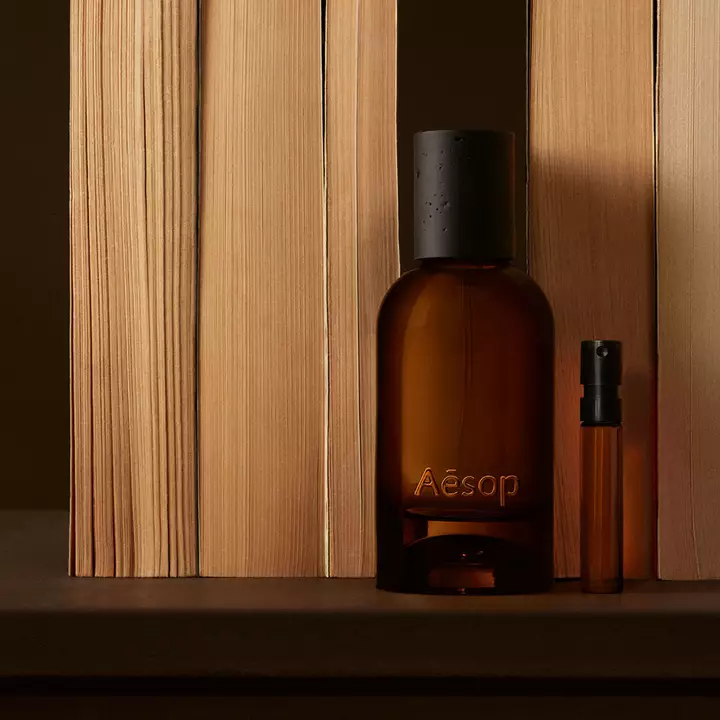 Bernsteinfarbene Aesop-Flasche und schmales Flaschchen zwischen vertikalen, hellen Holzbrettern, mit tiefbraunen Schatten und einer klaren, linearen Komposition.