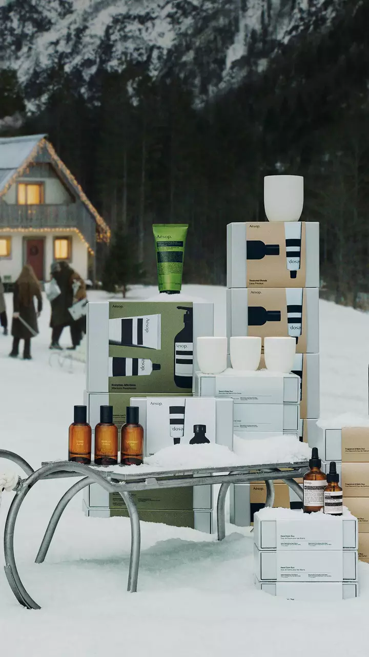Aesop-Schachteln, bernsteinfarbene Flaschen, farbige Tuben und weisse Gefasse sind im Schnee um einen Metallschlitten angeordnet, dahinter Menschen vor einem erleuchteten Haus.