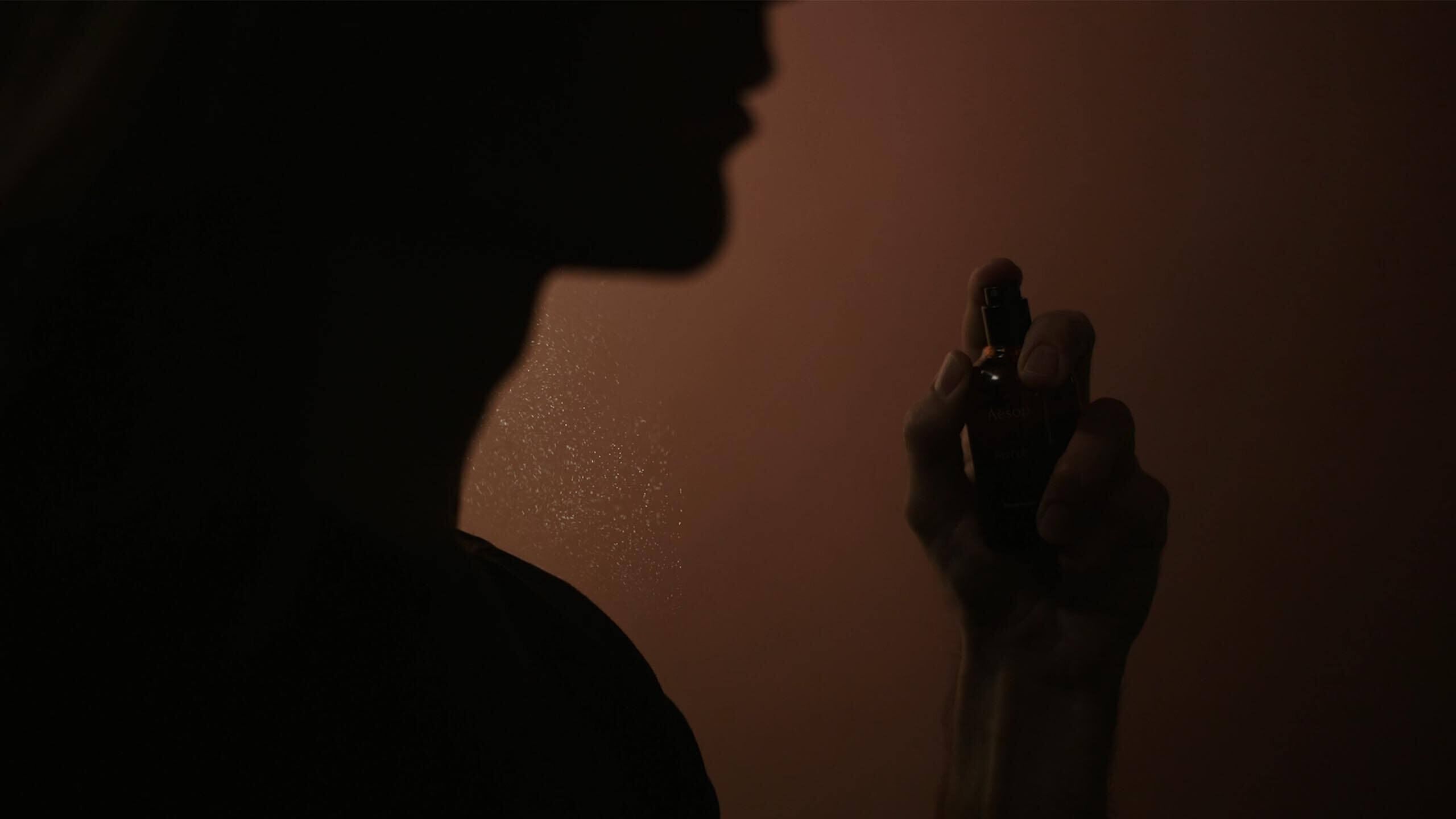 Silhouette einer Person, die ein kleines Duftflaschchen vor dem Gesicht vor einem warmen bernsteinfarbenen Hintergrund halt.