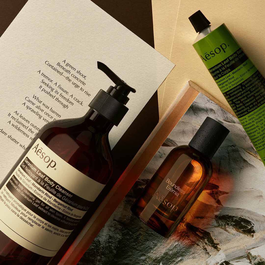 Aesop Boundless Eremia Flakon mit Pumpspender und grunem Schlauch, arrangiert auf Papieren und einem Steinbild.