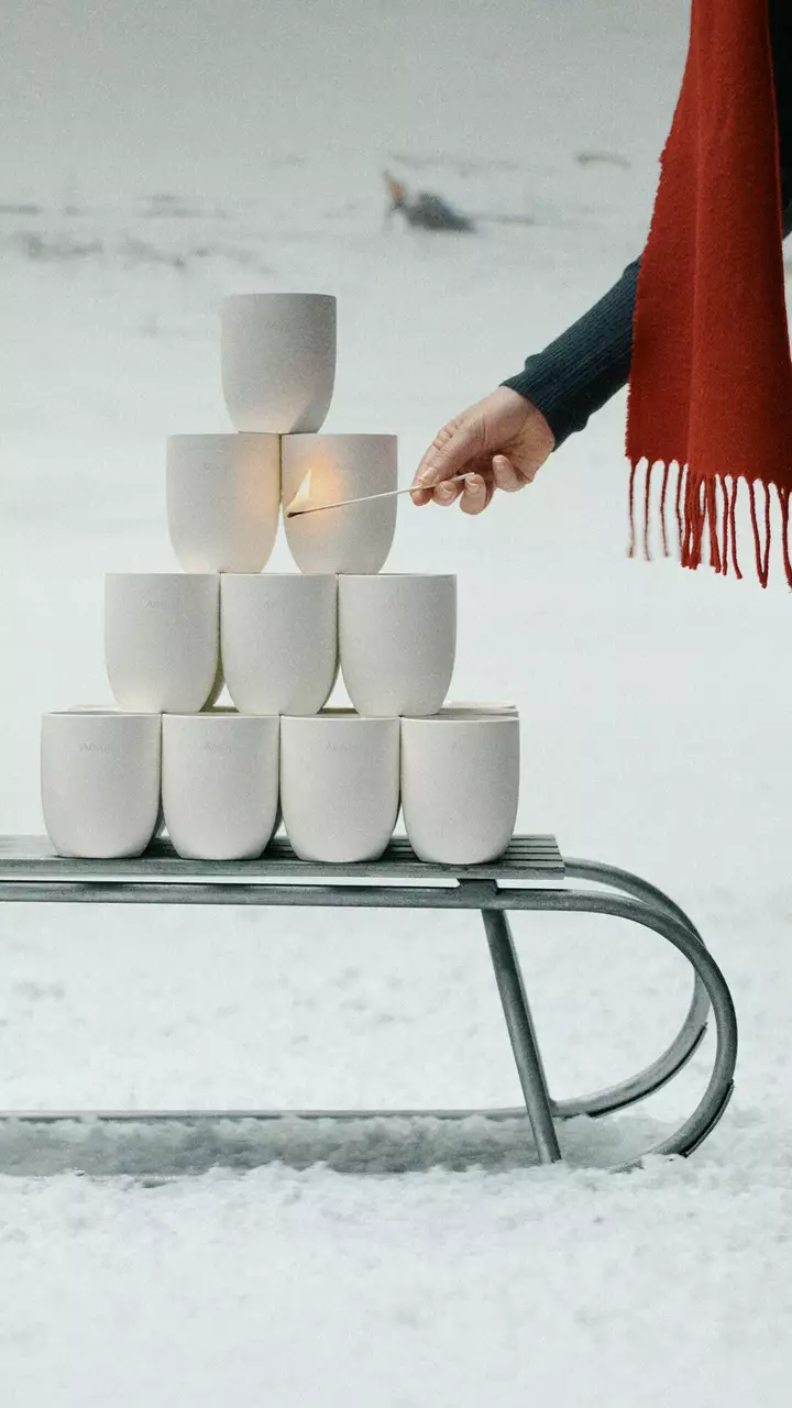 Eine Hand halt ein brennendes Streichholz neben weißen Gefaßen, die auf einem Metallschlitten im Schnee gestapelt sind; rechts liegt ein roter Schal mit Fransen.